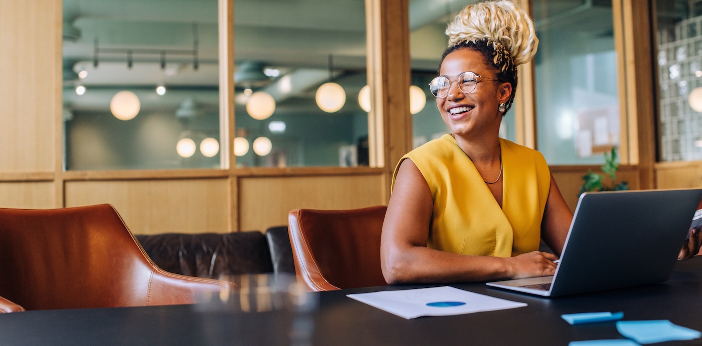 Image 2 Cheerful young black woman wearing glasses, using a laptop in a bright office setting. She radiates positivity and focus, surrounded by modern interior elements reflecting a professional environment.
