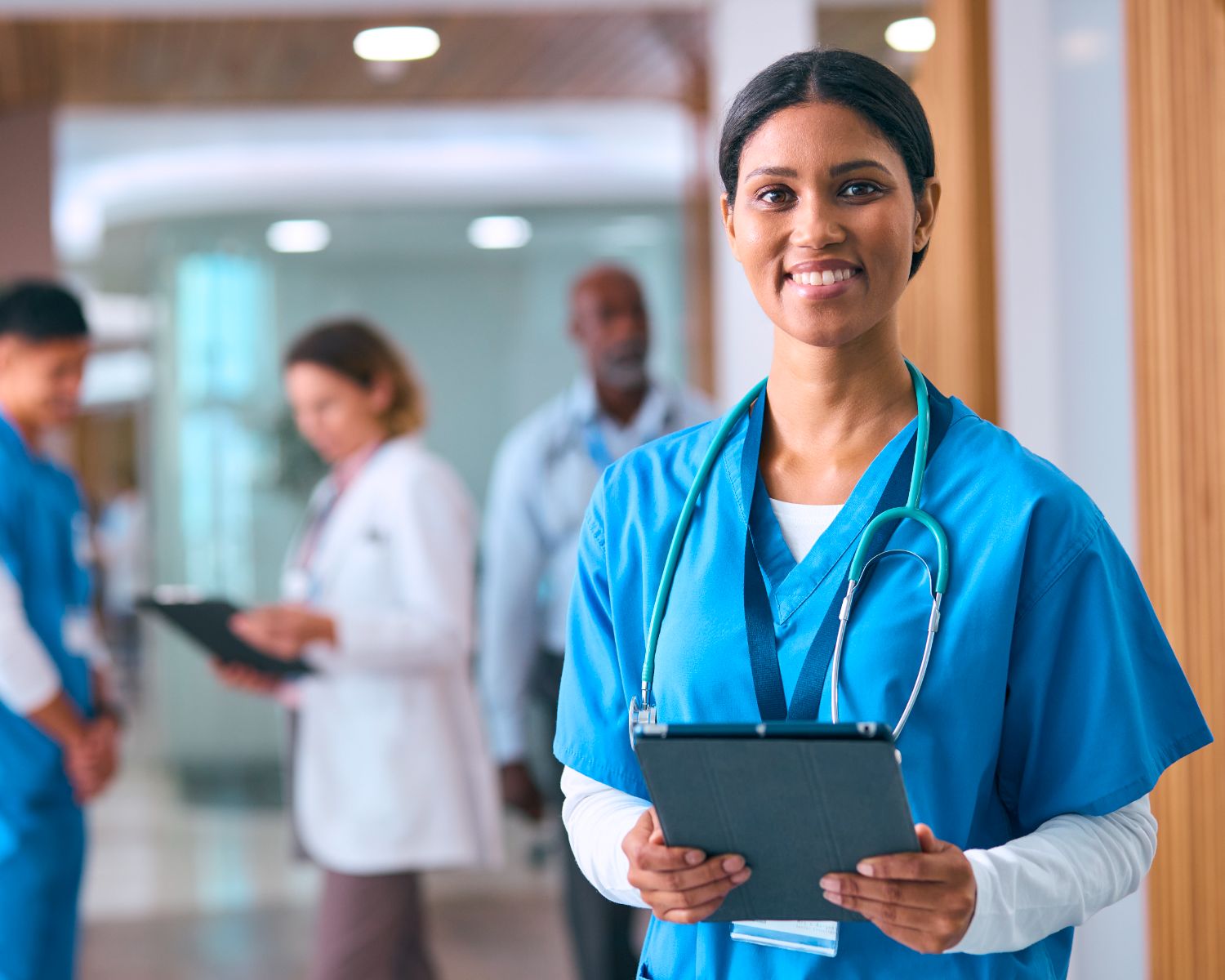 A smiling nurse in blue scrubs holding a tablet in a hospital hallway