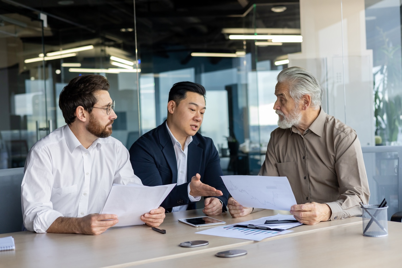 Image 2 Three diverse business people talking together sitting at a meeting desk inside office, businessmen in business suits discussing with boss business owner about financial indicators, achievements.
